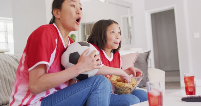 Mother and daughter sharing an exciting moment in living room while watching a soccer game on TV. The mother holds a soccer ball, and both are wearing red jerseys with snacks and drinks on the table. Ideal for themes of family bonding, sports enthusiasm, and cozy home settings.