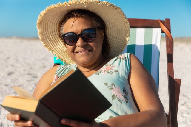 Senior African American woman enjoying a sunny day at the beach while reading a book. She is sitting on a folding chair, wearing sunglasses and a wide-brimmed hat, embodying relaxation and leisure. Perfect for use in articles or advertisements related to retirement, active lifestyles, summer vacations, and hobbies.