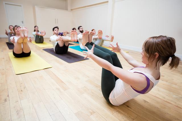 Group of individuals practicing boat pose during a yoga class in a fitness studio. Ideal for promoting fitness, wellness, and healthy lifestyle content. Useful for illustrating group exercise, flexibility training, and indoor workout sessions.