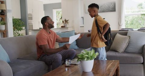 African American Father Celebrating Son's School Success with Backpack in Bright Living Room