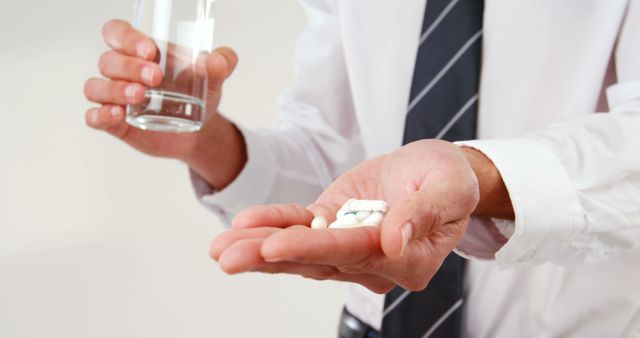A middle-aged Caucasian businessman is holding medication in one hand and a glass of water in the other, with copy space. His actions suggest he is about to take the pills, as part of a daily health routine or to address a specific ailment.