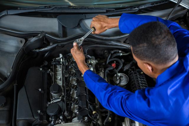 Mechanic in blue uniform working on car engine in auto workshop. Useful for content on car maintenance, vehicle repair services, and automotive industry. Ideal for illustrating the work of a car mechanic or auto technician.