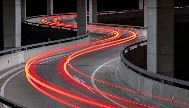 Spiral parking ramp showing glowing red taillight trails and sweeping concrete curves at night