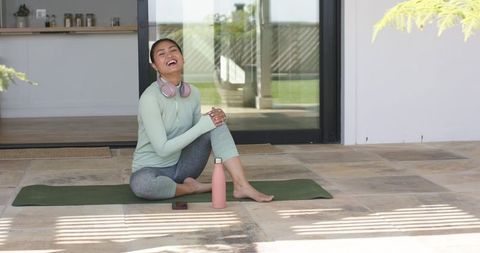 Asian woman practicing yoga on sunlit patio, wearing headphones, enjoying wellness