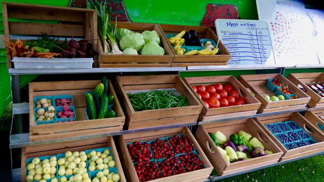 Outdoor farmers market display showcases fresh vegetables arranged in wooden crates. Price list for produce visible. Vibrant colors of tomatoes, peppers, cucumbers, green beans, leafy greens. Ideal for themes related to nutrition, healthy eating, organic farming, sustainable living.