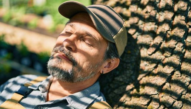 Hispanic gardener resting against tree with sunlit dappled shadows, cap and apron
