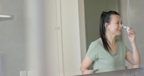 Asian woman using electric nasal care device at vanity mirror, minimalist bathroom routine