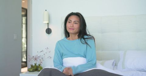 Asian woman sitting on bed in modern minimalist bedroom wearing light blue top, embracing calm