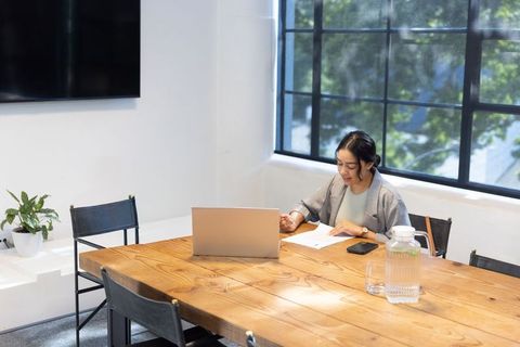 Asian businesswoman reviewing documents with laptop in minimalist sunlit conference room