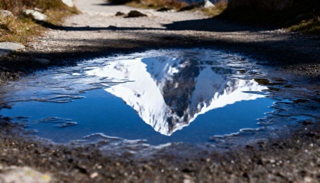 Snow-capped peak reflecting in puddle on alpine trail with blue sky, ice and gravel