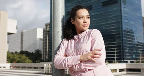 Young woman leaning on pole wearing pink hoodie gazing at city skyline on rooftop