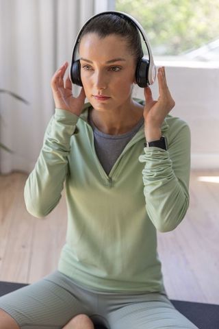 Woman adjusting over-ear headphones while meditating on mat in sunlit minimalist home