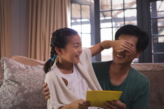 Daughter giving greeting card to father in living room, both smiling and enjoying the moment. Perfect for use in family-oriented advertisements, parenting blogs, greeting card promotions, and articles about family bonding and celebrations.