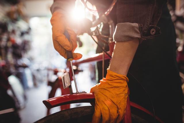 Mechanic wearing orange gloves is repairing a bicycle in a workshop with sunlight streaming in the background. Perfect for advertisements or articles related to bicycle maintenance, repair services, mechanical work, or professional cycling. Can also be used for blogs about DIY bike repair tips or for promoting bike shops.