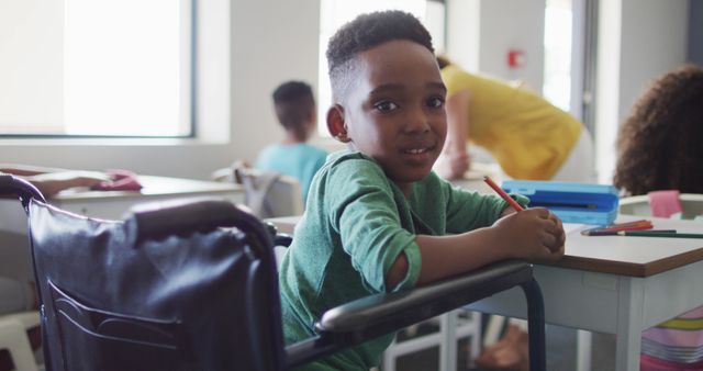 Young boy in a wheelchair is smiling at his desk in a classroom, engaging in an inclusive learning environment. Great for illustrating themes such as education, diversity, inclusion, accessibility, and student life. Ideal for use in educational materials, website content, and articles about inclusive education and accessibility.
