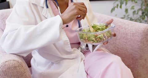 Clinician eating salad on pastel sofa during break wearing pink scrubs and lab coat