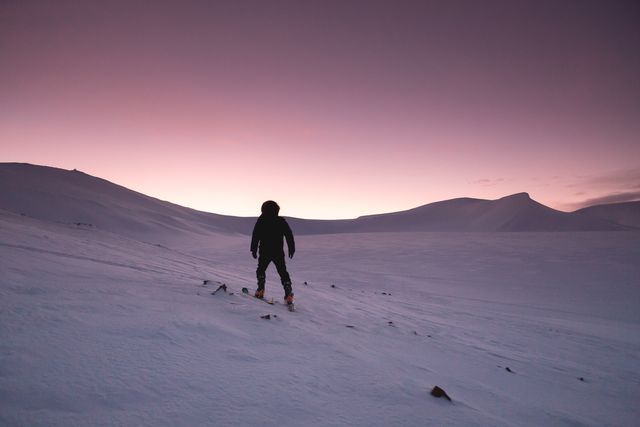 Person wearing winter gear is hiking through snow-covered mountainous terrain during sunset. This image can be used to illustrate themes of adventure, exploration, solitude, winter activities, and the beauty of winter landscapes. Ideal for travel blogs, outdoor adventure articles, and winter sports promotions.