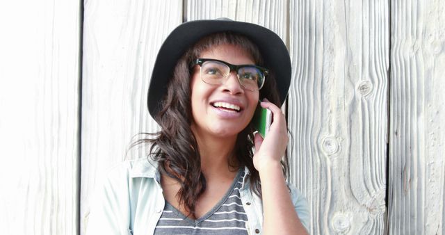 Woman wearing glasses and a stylish hat smiling while talking on her smartphone, against a wooden background. Useful for themes related to modern communication, fashion, happiness, and casual lifestyle.