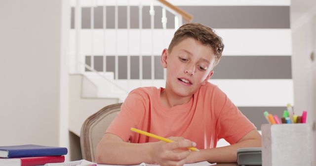 This image depicts a young boy in an orange shirt intensely focused on his homework while sitting at home. The background features a staircase and a striped wall, and various school supplies like colored pens are seen around his workspace. This can be used for promoting education services, online learning platforms, school programs, or articles about the importance of study habits in children.