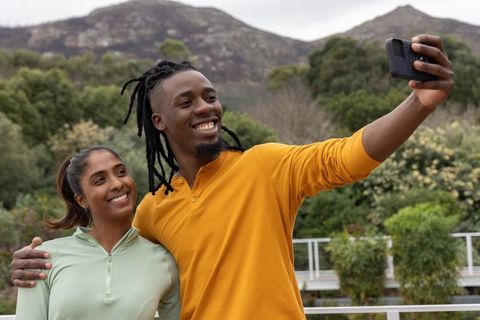 Diverse couple taking selfie on outdoor deck with mountain backdrop smiling and embracing