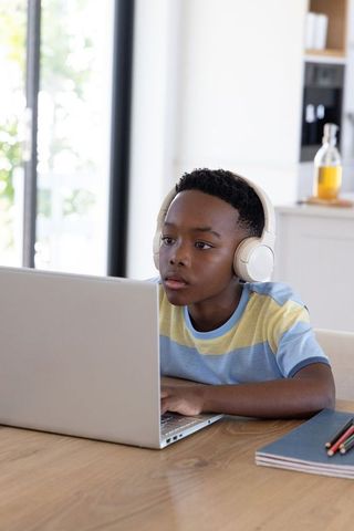 African american boy studying on laptop wearing headphones in bright modern kitchen