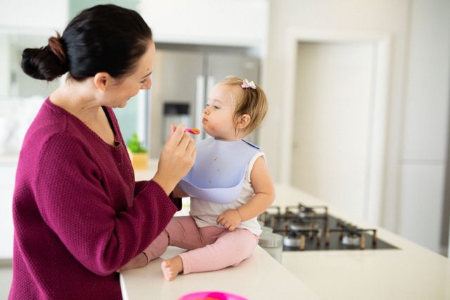 Mother feeding baby daughter in modern kitchen. They are sitting on counter top, enjoying mealtime together. Ideal for themes related to parenting, family life, childcare, and home activities. Can be used in articles, blogs, and advertisements focused on family, health, and nutrition.