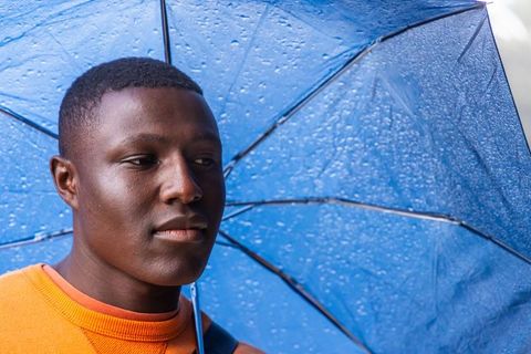 African american man holding wet blue umbrella on rainy urban street, contemplative closeup