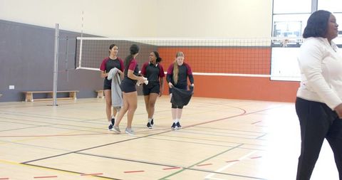 Diverse high school girls volleyball team walking across indoor gym court holding towels