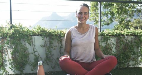Smiling woman sitting cross-legged on sunlit balcony wearing red leggings and tank top