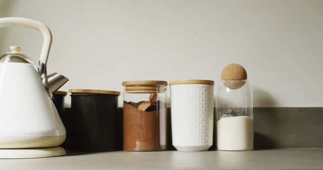 Modern kitchen shows minimalist design with white kettle placed on kitchen counter next to storage jars filled with ingredients such as brown sugar and white sugar, all having wooden lids. Perfect for home organization websites, minimalist and contemporary kitchen design blogs, and online furniture shops showcasing kitchen accessories.