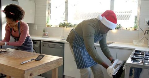 African american couple baking holiday cookies in sunlit kitchen, man wearing santa hat