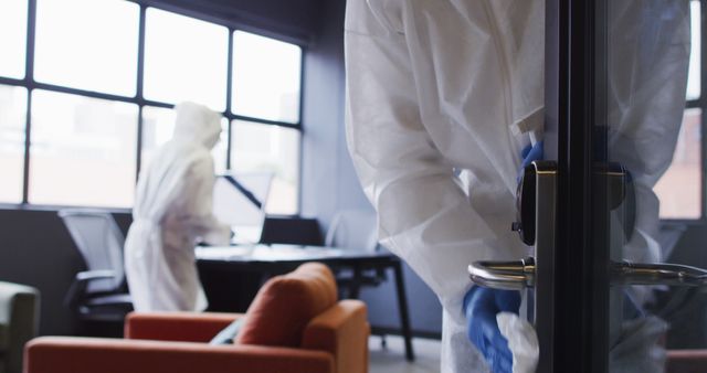 This photo depicts two cleaners in full protective suits and gloves disinfecting an office space. The foreground shows one cleaner working on a door handle, while another cleaner is visible in the background wiping down a desk. This image is ideal for use in articles, advertisements, or campaigns focused on health and safety measures, workplace hygiene, pandemic precautions, and cleaning services.