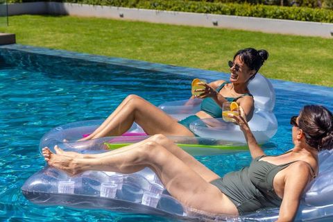 Diverse women relaxing on clear inflatable floats sipping cocktails in sunny backyard pool