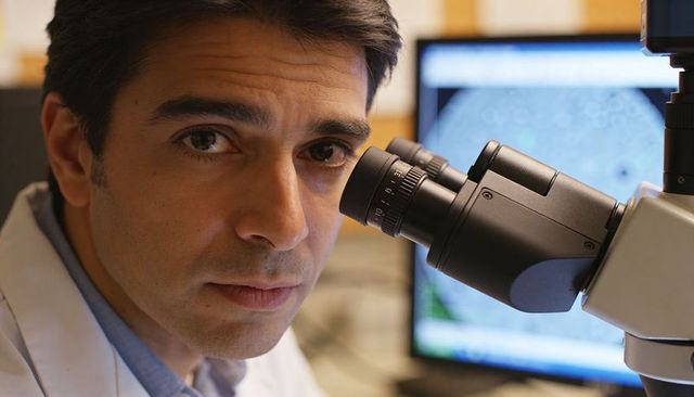 Focused scientist examining samples through microscope with computer monitor in lab