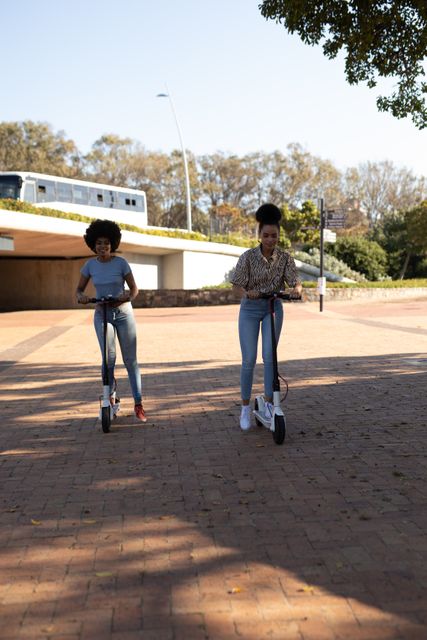 Front view of two happy biracial twin sisters enjoying free time together in a city on a sunny day, riding electric scooters in an urban park.