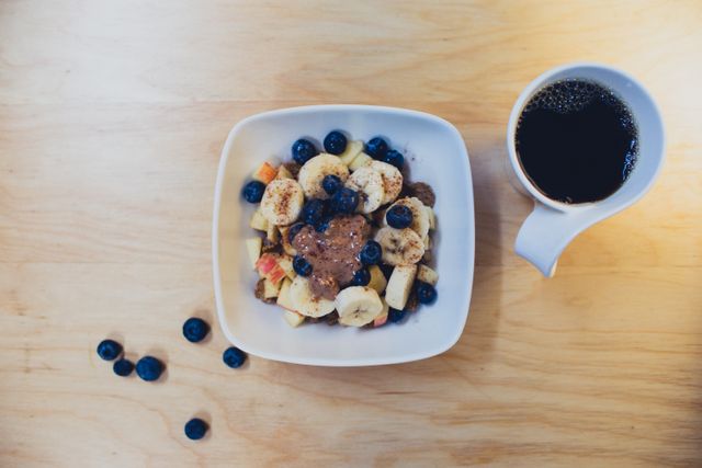Colorful breakfast bowl with blueberries, sliced bananas, apples, and nut butter accompanied by a cup of black coffee on a light wooden table. Great for promoting healthy eating, nutritious breakfast options, and balanced diets. Depicts a fresh, energetic start to the day.