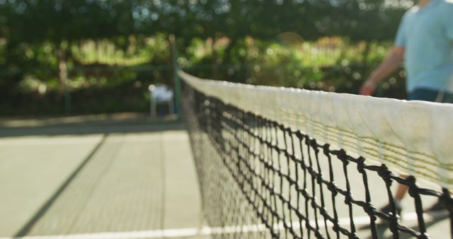 The net divides a tennis court on a sunny day, foreground focused while the background is blurred. The image emphasizes the structured net and creates depth with a player in the distance. Ideal for use in articles about tennis, sports equipment, outdoor recreation, or focusing on the details of sports settings.