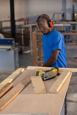 Craftsman aligning wooden plank on workbench holding nail gun wearing safety gear