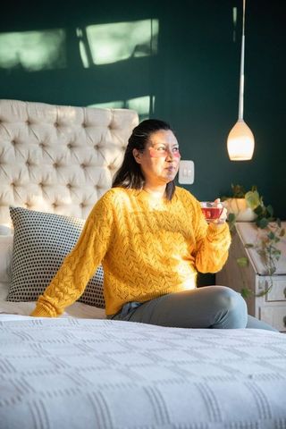Sunlit woman sitting on bed wearing yellow sweater holding glass cup of tea, cozy morning