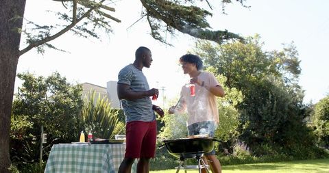 Diverse friends grilling at charcoal barbecue in sunny backyard gathering with drinks