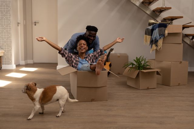 This image captures a joyful moment of a young African American couple enjoying their moving day. The man is playfully pushing his girlfriend who is sitting in a cardboard box, while their dog walks around. Ideal for use in articles or advertisements related to moving services, homeownership, relationships, and lifestyle blogs.