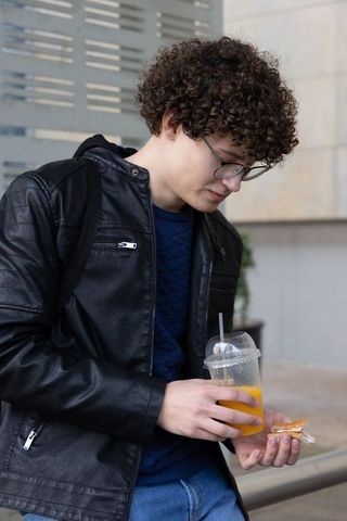 Curly-haired teen holding orange juice and sandwich in leather jacket, urban streetwear lifestyle