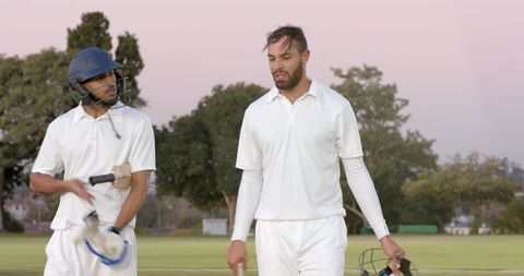 Diverse cricket teammates walking off grassy field at dusk carrying bats and helmets