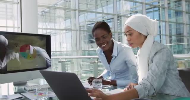 Two businesswomen engaging with digital technology, one wearing a hijab, depict diversity and teamwork. Modern office environment with natural light and professionalism portrayed. Suitable for themes of business communication, multicultural workplaces, and remote collaboration.