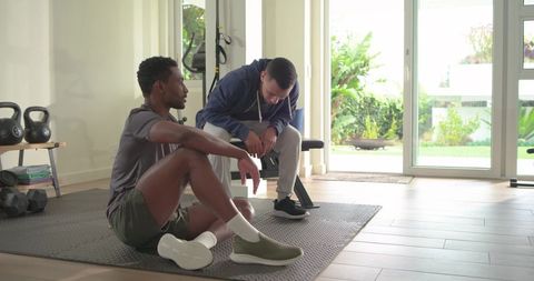 Multiracial men resting during home workout with personal trainer on mat and garden view