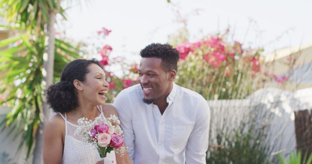 Shows a cheerful couple smiling and laughing while holding a bouquet of flowers in a summer garden. Perfect for promoting romance, love, and wedding themes. Suitable for use in wedding invitations, engagement announcements, lifestyle blogs, and relationship advice websites.