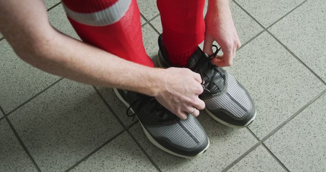 Image shows an athlete tying the laces on sport shoes while wearing red socks, standing on a tiled floor. Perfect for illustrating themes of athletic preparation, fitness routines, footwear styling for sports, and pre-training rituals.
