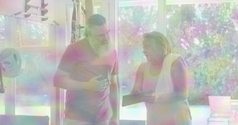 Senior female trainer coaching mature man in sunlit home gym with tablet and dumbbells
