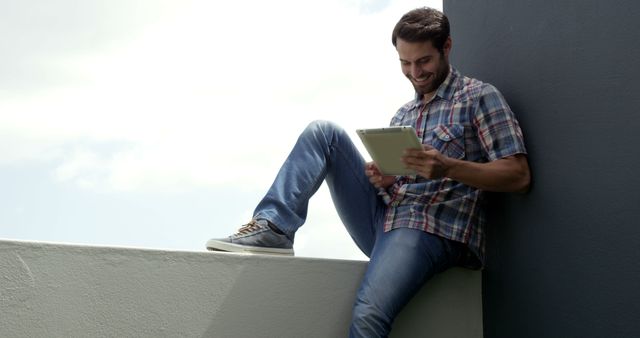 Young man sits on outdoor ledge engaging with tablet. Suitable for illustrating relaxation, leisure activities, use of technology in everyday life, and modern digital lifestyle.