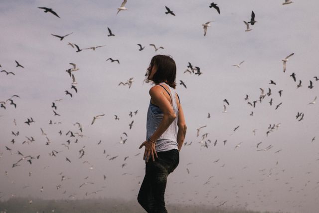 A woman standing outdoors surrounded by a flock of seagulls in a foggy environment. She is gazing upwards, creating a serene and contemplative mood. Ideal for themes of freedom, solitude, harmony with nature, wanderlust, and inspiration in outdoor adventure blogs, travel promotions, and mindfulness content.